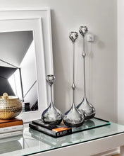 Load image into Gallery viewer, Three candle holderson a glass table with a mirror and books in the background. Lumea Silver Candle Holders by Allthingscurated feature a striking trio of sculptural pieces that blend contemporary design with timeless elegance. Each holder features a distinctive water droplet base that anchors a sleek, minimalist silhouette, creating an art deco-inspired statement on its own. this set makes an exceptional wedding gift for couples seeking sophisticated home décor that transcends occasion.
