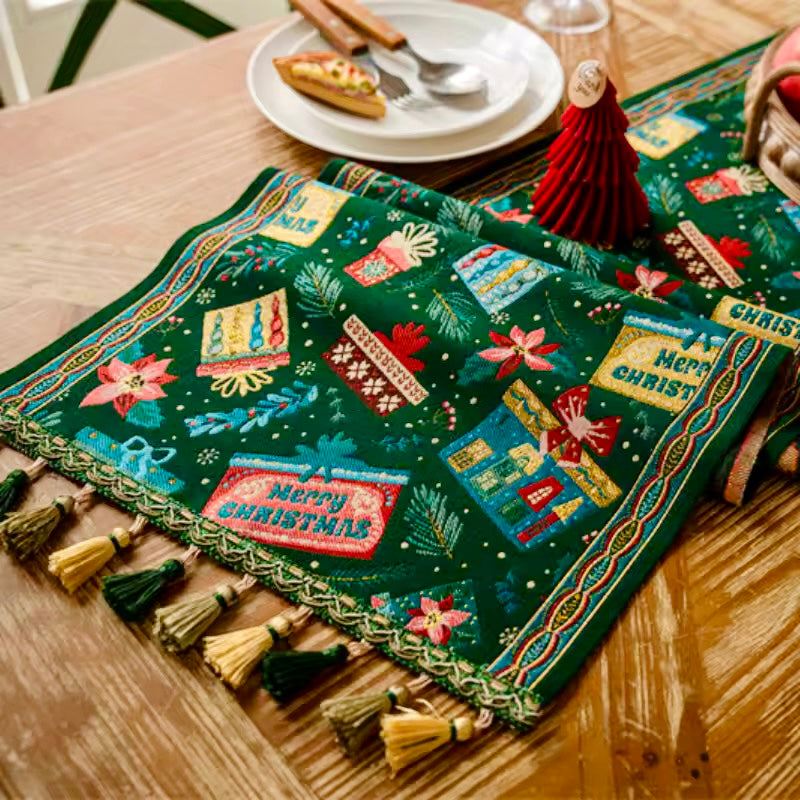 Decorative Christmas table runner with tassels on a wooden table. Elevate your holiday entertaining with this Merry Christmas Jacquard Table Runner by Allthingscurated. Inspired by the joy of gift-giving, this table runner features an array of gift box motifs rendered in rich jewel tones, this piece showcases beautiful embroidery complemented by pretty tassels. Perfect for elevating your holiday tablescape, making every feast and gathering truly memorable. 