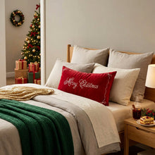 Load image into Gallery viewer, A bed with neutral linens, a green knit throw, a gold sequined blanket, and a red velvet Merry Christmas pillow, set against a decorated tree that offers inspiration for your holiday home decor.
