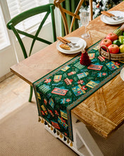 Load image into Gallery viewer, Decorative table runner with festive design on a wooden table. Elevate your holiday entertaining with this Merry Christmas Jacquard Table Runner by Allthingscurated. Inspired by the joy of gift-giving, this table runner features an array of gift box motifs rendered in rich jewel tones, this piece showcases beautiful embroidery complemented by pretty tassels. Perfect for elevating your holiday tablescape, making every feast and gathering truly memorable. 
