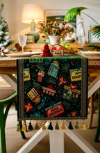 Load image into Gallery viewer, Decorative table runner with festive designs on a Christmas-themed table. Elevate your holiday entertaining with this Merry Christmas Jacquard Table Runner by Allthingscurated. Inspired by the joy of gift-giving, this table runner features an array of gift box motifs rendered in rich jewel tones, this piece showcases beautiful embroidery complemented by pretty tassels. Perfect for elevating your holiday tablescape, making every feast and gathering truly memorable.
