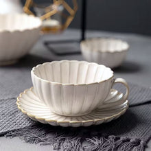 Load image into Gallery viewer, Off-white ceramic cup and saucer on a textured surface with blurred background. Japanese Chrysanthemum Cup and Saucer by Allthingscurated is inspired by the flower’s perfect symmetry and stunning beauty in the Japanese culture. Crafted from porcelain, it’s a beautiful set to entertain friends and family for afternoon teas at home.
