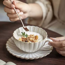 Load image into Gallery viewer, Person holding a spoon over a bowl of yogurt with granola on a wooden table. Japanese Chrysanthemum Cup and Saucer by Allthingscurated is inspired by the flower’s perfect symmetry and stunning beauty in the Japanese culture. Crafted from porcelain, it’s a beautiful set to entertain friends and family for afternoon teas at home.
