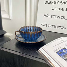 Load image into Gallery viewer, Blue ceramic cup and saucer on a dark surface with a magazine open to a page in the background. Japanese Chrysanthemum Cup and Saucer by Allthingscurated is inspired by the flower’s perfect symmetry and stunning beauty in the Japanese culture. Crafted from porcelain, it’s a beautiful set to entertain friends and family for afternoon teas at home.

