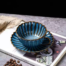 Load image into Gallery viewer, Blue ceramic cup set on a magazine with coffee beans in the background. Japanese Chrysanthemum Cup and Saucer by Allthingscurated is inspired by the flower’s perfect symmetry and stunning beauty in the Japanese culture. Crafted from porcelain, it’s a beautiful set to entertain friends and family for afternoon teas at home.
