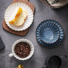 Load image into Gallery viewer, Ceramic dishes with coffee beans, slices of bread, and an empty blue cup saucer set on a gray surface. Japanese Chrysanthemum Cup and Saucer by Allthingscurated is inspired by the flower’s perfect symmetry and stunning beauty in the Japanese culture. Crafted from porcelain, it’s a beautiful set to entertain friends and family for afternoon teas at home.
