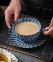 Load image into Gallery viewer, Blue ceramic cup set with a decorative interior filled with tea, held by a person. Japanese Chrysanthemum Cup and Saucer by Allthingscurated is inspired by the flower’s perfect symmetry and stunning beauty in the Japanese culture. Crafted from porcelain, it’s a beautiful set to entertain friends and family for afternoon teas at home.
