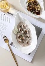 Load image into Gallery viewer, Two white oyster shape with seafood cuisines on a textured surface with a magazine underneath. Oyster Shape Serving Dish by Allthingscurated is elegantly crafted in pristine white ceramic to mirror the contours and texture of the elegant oyster. The striking serve ware comes in multiple sizes for plating appetizers or main courses.
