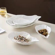 Load image into Gallery viewer, Set of white oyster shape ceramic dishes on a light surface with a glass of water in the background. Oyster Shape Serving Dish by Allthingscurated is elegantly crafted in pristine white ceramic to mirror the contours and texture of the elegant oyster. The striking serve ware comes in multiple sizes for plating appetizers or main courses.
