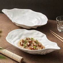 Load image into Gallery viewer, Two white oyster-shaped ceramic dishes on a brown surface with a glass and chopsticks. Oyster Shape Serving Dish by Allthingscurated is elegantly crafted in pristine white ceramic to mirror the contours and texture of the elegant oyster. The striking serve ware comes in multiple sizes for plating appetizers or main courses.
