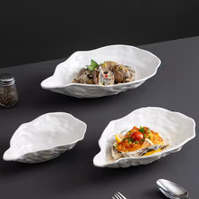 Load image into Gallery viewer, Set of white oyster-shaped ceramic dishes with food on a dark surface. Oyster Shape Serving Dish by Allthingscurated is elegantly crafted in pristine white ceramic to mirror the contours and texture of the elegant oyster. The striking serve ware comes in multiple sizes for plating appetizers or main courses.
