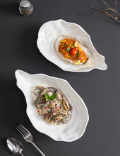 Load image into Gallery viewer, Two white oyster shape ceramic dishes with oyster dishes on a dark surface. Oyster Shape Serving Dish by Allthingscurated is elegantly crafted in pristine white ceramic to mirror the contours and texture of the elegant oyster. The striking serve ware comes in multiple sizes for plating appetizers or main courses.
