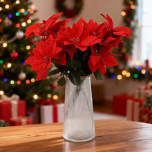 Load image into Gallery viewer, Bouquet of red poinsettias in a clear vase on a wooden table with a festive background. Bring the true spirit of the holidays into your home with this bundle of 5 classic red Faux Poinsettia by Allthingscurated. These timeless blooms complement any festive palette and instantly evokes Christmas cheer.
