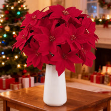 Load image into Gallery viewer, Red poinsettia flowers in a white vase on a wooden table with a Christmas tree in the background. Bring the true spirit of the holidays into your home with this bundle of 5 classic red Faux Poinsettia by Allthingscurated. These timeless blooms complement any festive palette and instantly evokes Christmas cheer.
