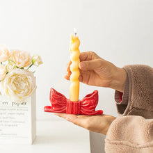 Load image into Gallery viewer, Hand holding a twisted candle in a red bow candle holder with flowers in the background. Fashion Bow Candle Holder by Allthingscurated spots a distinctive knot design, beautiful yet practical. Perfect for wedding celebrations, home styling, or to infuse warmth and romance into any living space. Available in three colors and 2 sizes. 
