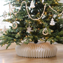 Load image into Gallery viewer, Decorated Christmas tree with white ornaments and a paper collar on a wooden floor. Transform your holiday setup with the striking Honeycomb Christmas Tree Collar by Allthingscurated. Crafted from premium paper with easy setup, its distinctive pleated design delivers sophisticated visual interest while concealing the tree base with understated elegance. The crips white finish works beautifully with any tree style.
