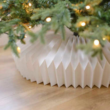 Load image into Gallery viewer, White paper collar under a Christmas tree with lights on a wooden floor. Transform your holiday setup with the striking Honeycomb Christmas Tree Collar by Allthingscurated. Crafted from premium paper with easy setup, its distinctive pleated design delivers sophisticated visual interest while concealing the tree base with understated elegance. The crips white finish works beautifully with any tree style.
