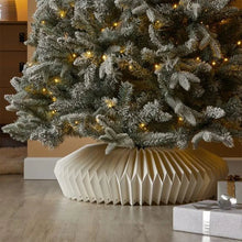 Load image into Gallery viewer, Decorated Christmas tree with lights and a white paper collar in a room setting. Transform your holiday setup with the striking Honeycomb Christmas Tree Collar by Allthingscurated. Crafted from premium paper with easy setup, its distinctive pleated design delivers sophisticated visual interest while concealing the tree base with understated elegance. The crips white finish works beautifully with any tree style.

