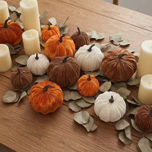 Load image into Gallery viewer, A decorative autumn centerpiece with multiple faux velvet pumpkins in orange, brown, and cream, arranged on a wooden table with eucalyptus leaves and white pillar candles.
