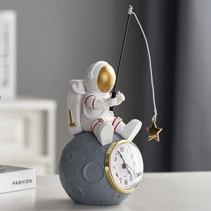  Allthingscurated Desk Clock featuring an astronaut sitting atop a moon rock fishing a star. Astronaut is dressed in a white space suit with a gold space mask, holding a fishing rod with a dangling gold star. Size of clock measuring height 24cm, width 10cm and length 10cm and weighing 415 gram.