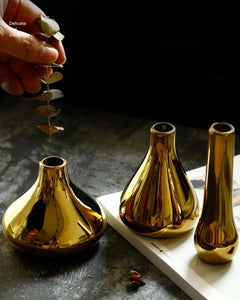 3 gold mini gold vases on a table top. A human hand is putting a strand of eucalyptus into a vase.